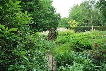 A Walter Bailey sculpture placed in a woodland wetland garden amongst strong foliage growth of Hosta, Aruncus and ferns.