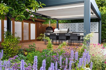 Modern pergola with outdoor kitchen and dining deck, surrounded by lush planting